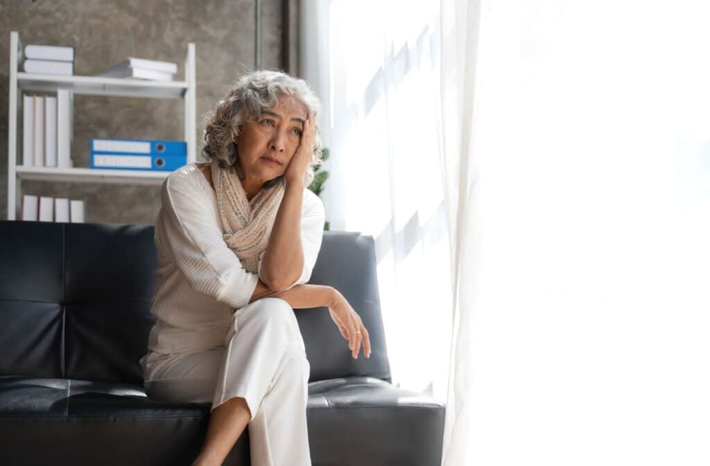 A senior holds a hand to their head as they look worryingly out of an open window while sitting on a black couch
