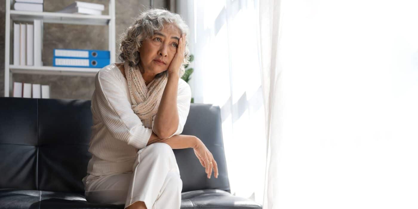 A senior holds a hand to their head as they look worryingly out of an open window while sitting on a black couch