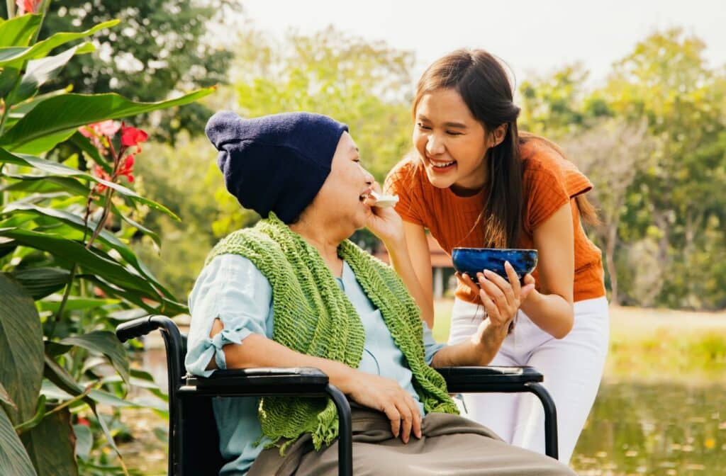An individual feeds their older parent soup while taking them out through a park on a walk with their wheelchair