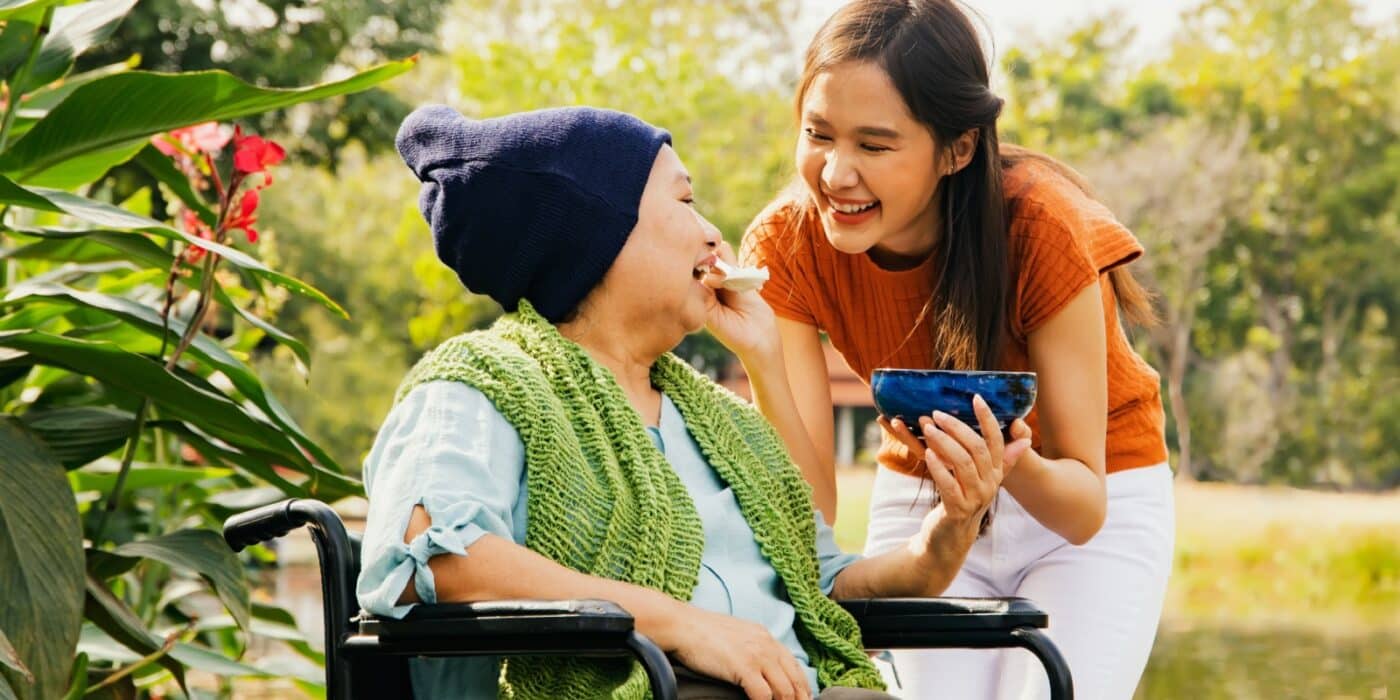 An individual feeds their older parent soup while taking them out through a park on a walk with their wheelchair