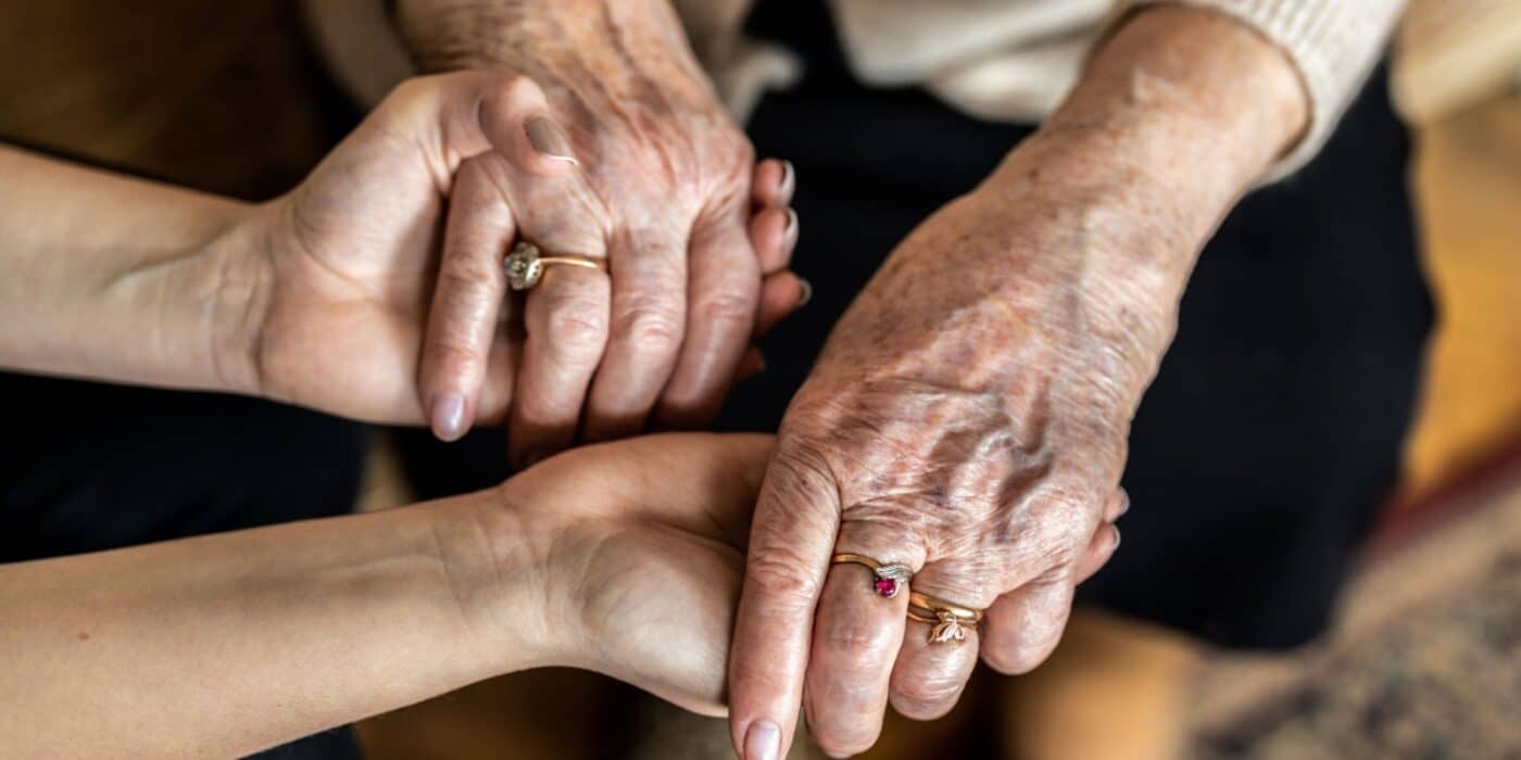 A middle-aged individual holds both of their senior parent’s hands to show their support