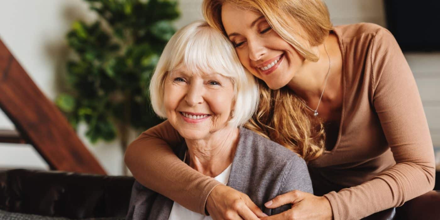 A senior mother being happily embraced by her loving daughter while seated on a couch.