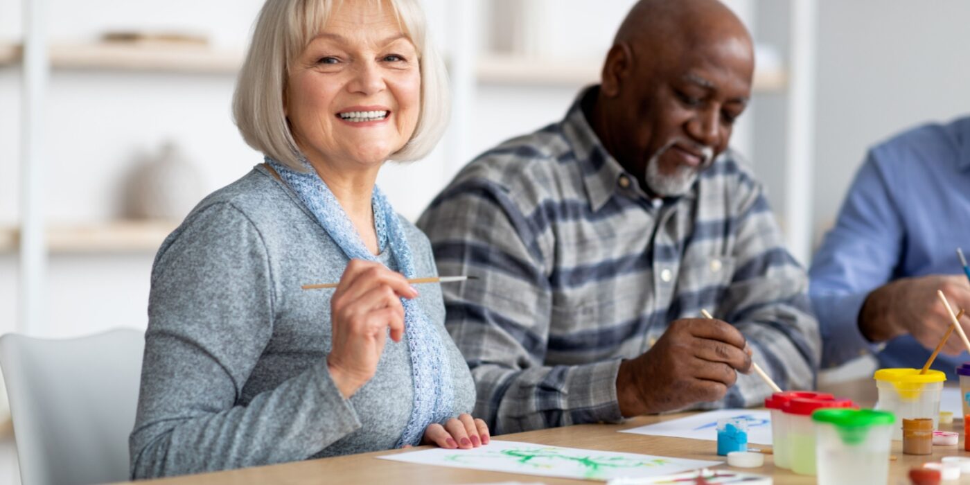 A senior in assisted living painting on a canvas while enjoying time with friends