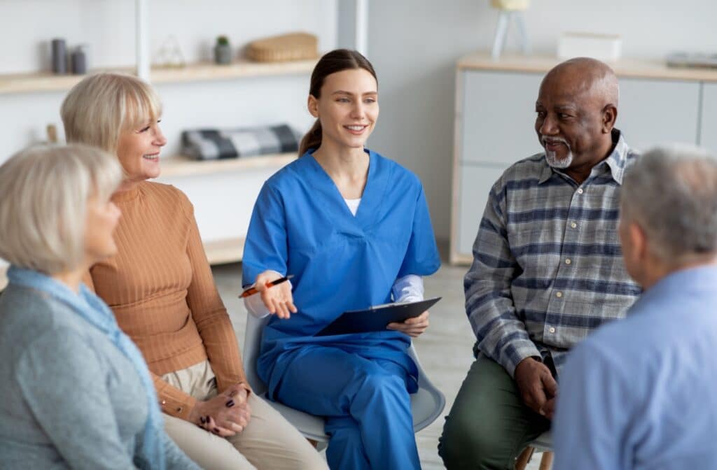 A nurse in blue scrubs sits in a circle with seniors while they explain some of the features of assisted living they can use