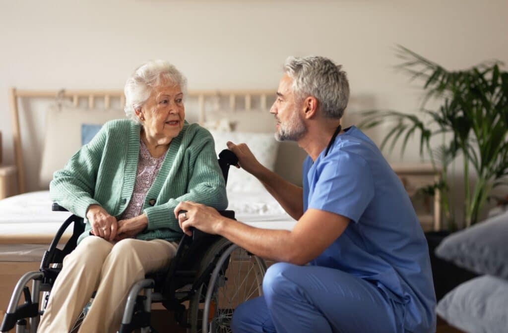 A senior woman in her wheelchair talking to her caregiver seated beside her in blue scrubs.