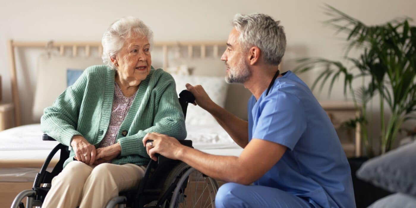 A senior woman in her wheelchair talking to her caregiver seated beside her in blue scrubs.