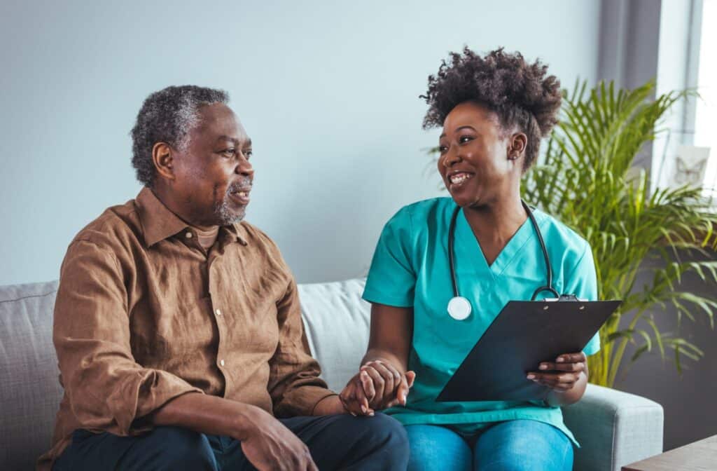 Smiling healthcare worker holding clipboard talks with older adult on couch in bright, home-like care setting