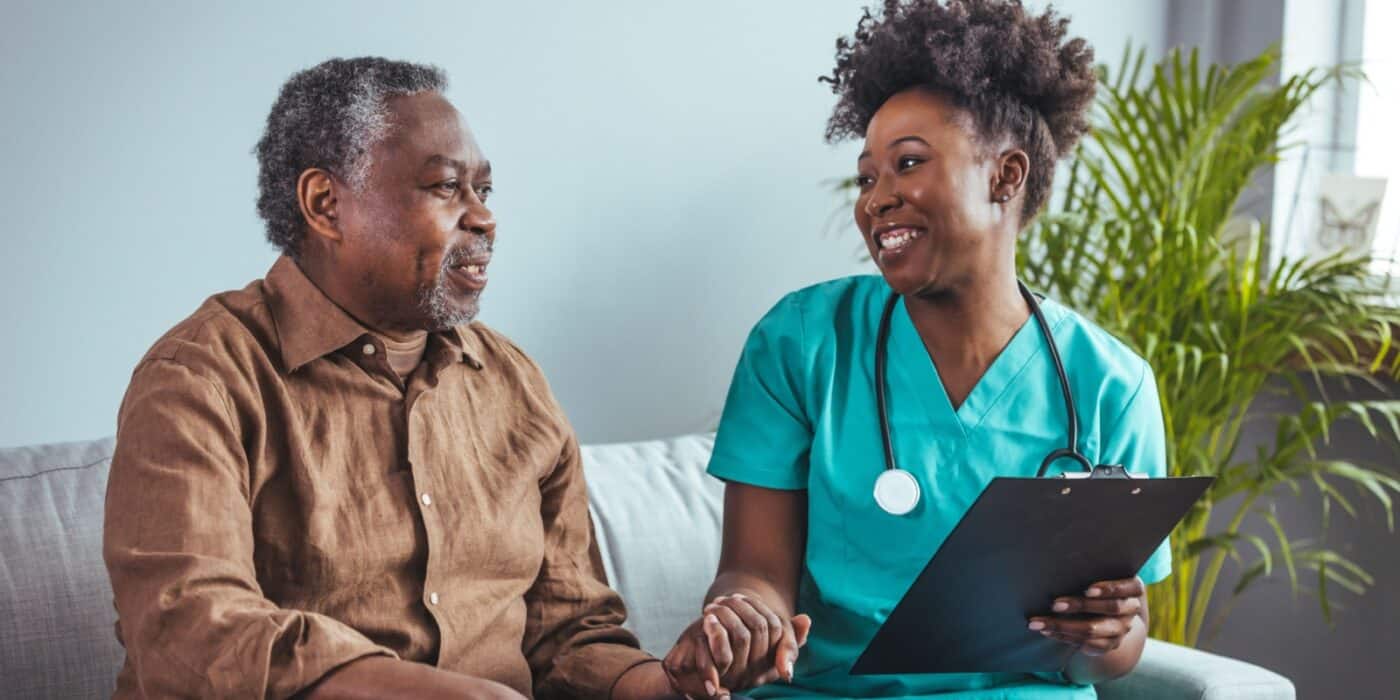 Smiling healthcare worker holding clipboard talks with older adult on couch in bright, home-like care setting