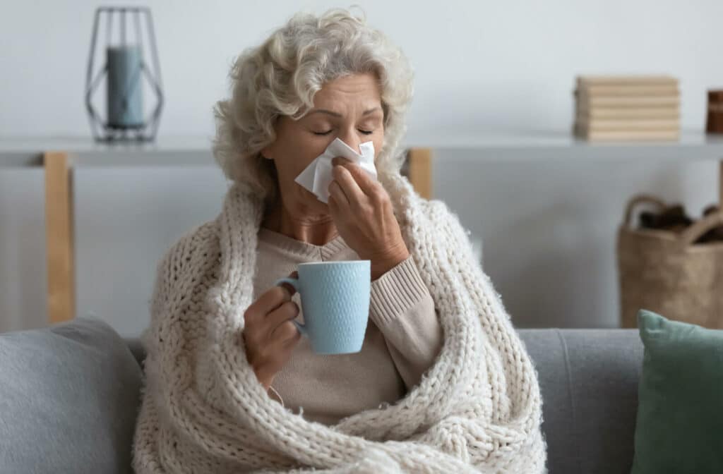 An older adult in a blanket blowing their nose and clasping a cup of tea to stop feeling so cold.