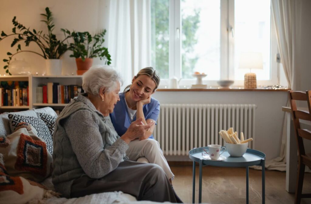 Caregiver and older adult smiling together on a couch in a cozy living room with pastries and tea on a small table nearby