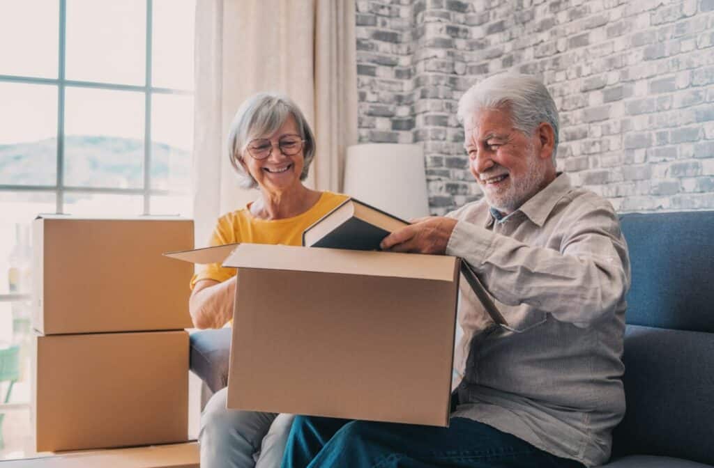 Smiling older adult couple sitting and unpacking books from moving boxes in a bright assisted living apartment