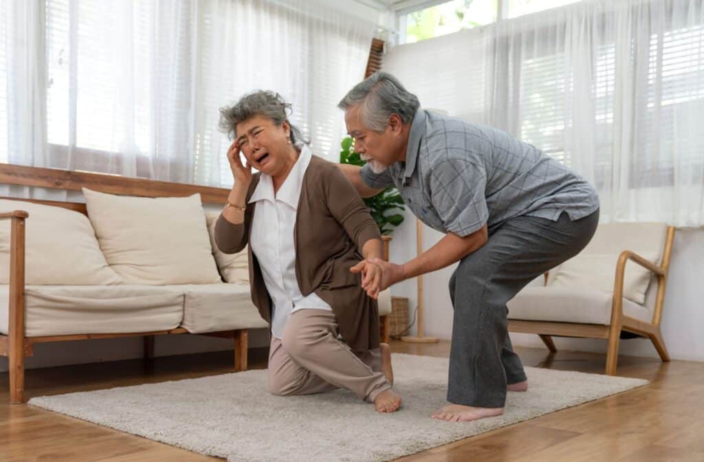Older adult holding head in pain while another assists them after a fall in a bright senior living room