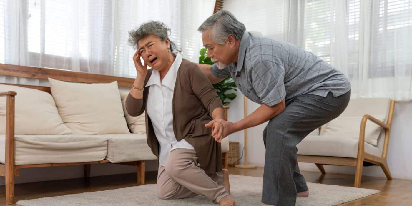 Older adult holding head in pain while another assists them after a fall in a bright senior living room
