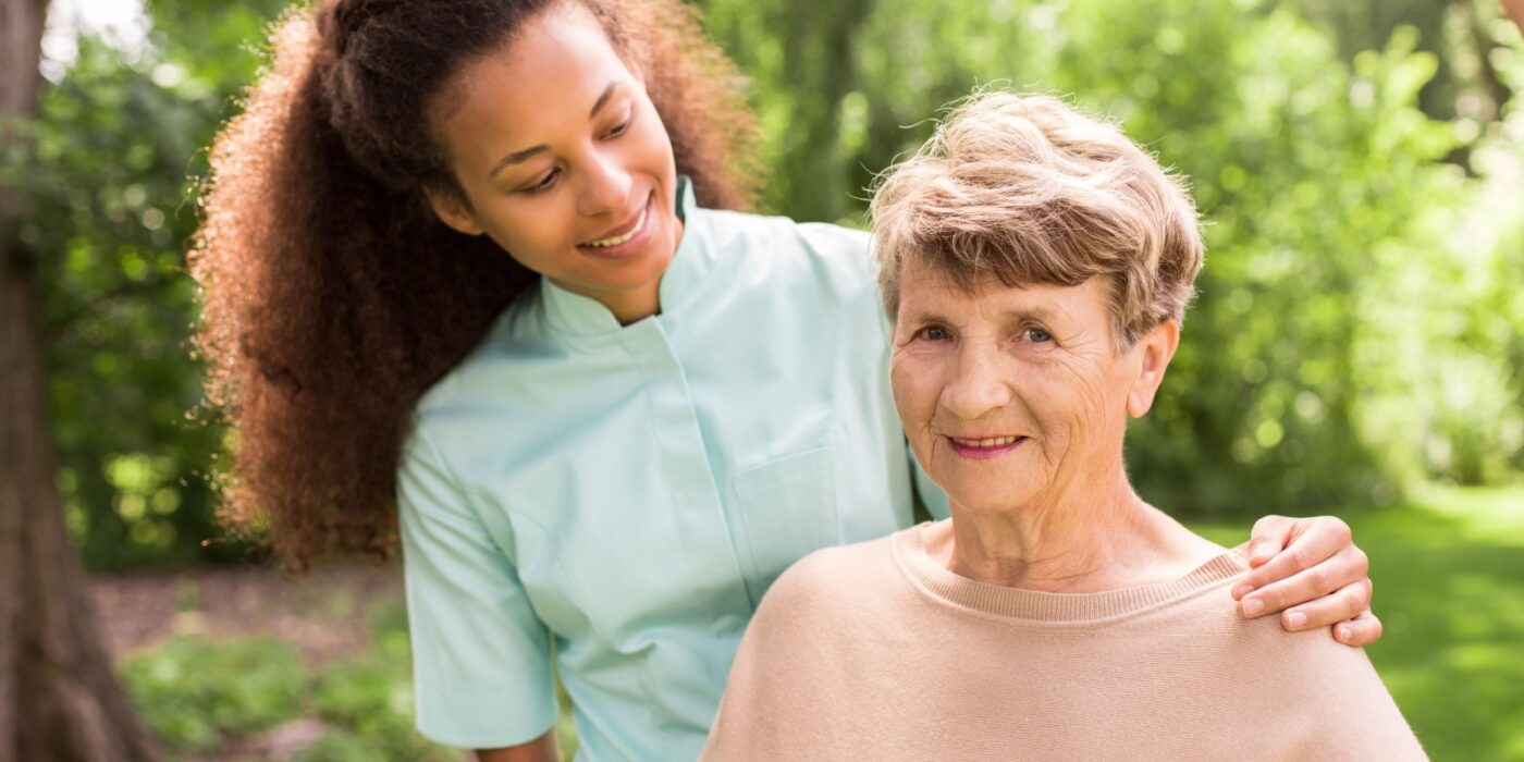 Caregiver supporting older adult outdoors surrounded by greenery