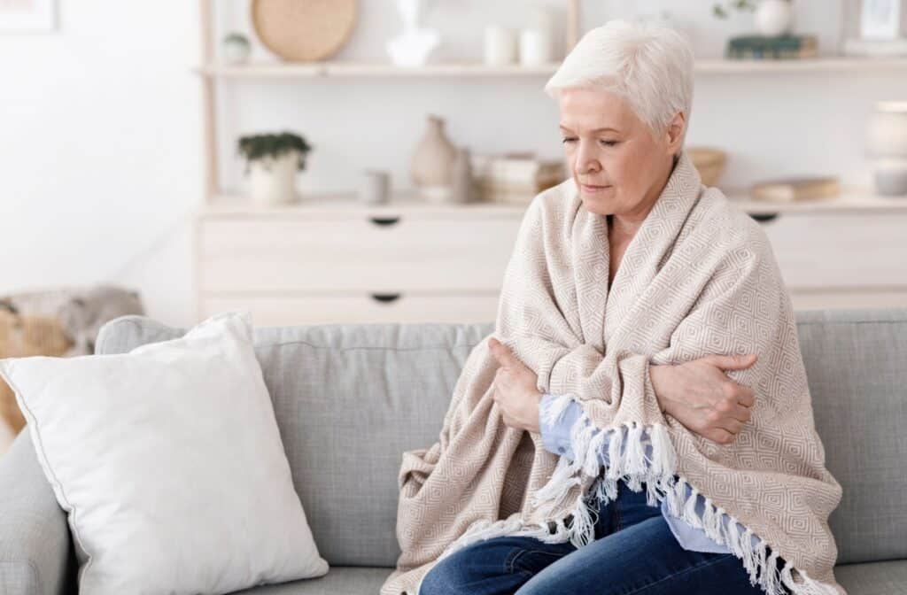 An older adult wrapped tightly in a blanket, sitting on their couch and shivering due to feeling cold.
