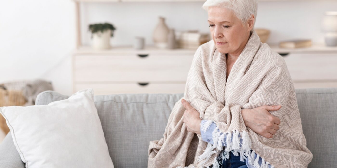 An older adult wrapped tightly in a blanket, sitting on their couch and shivering due to feeling cold.