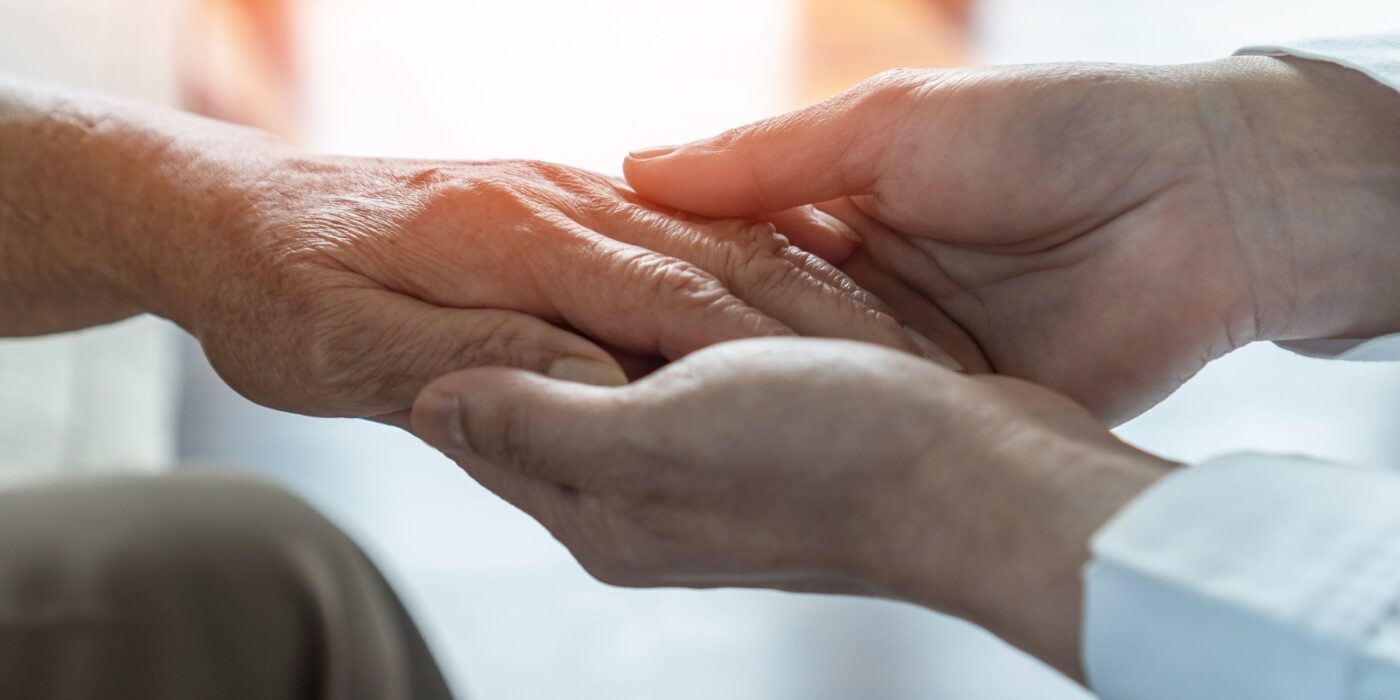 A caregiver takes the hand of an older adult with dementia in front of a sunlit window