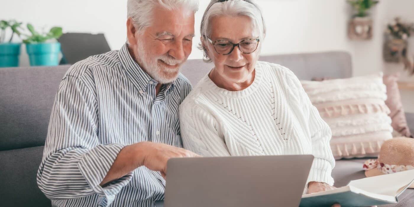An older adult grins while sitting on the couch beside their spouse and pointing at a laptop screen while researching assisted living