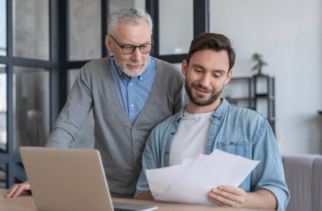 An older adult watches as their smiling adult child reviews paperwork while they budget together for assisted living