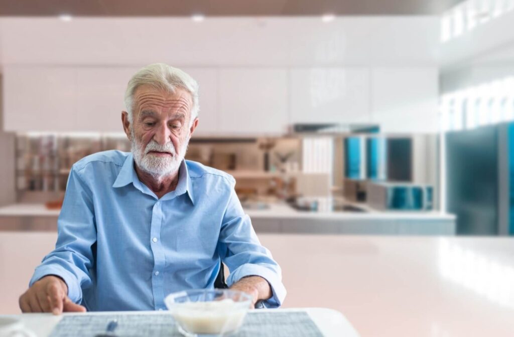 An older adult looks at a bowl of yogurt uninterestedly while sitting alone in their kitchen.