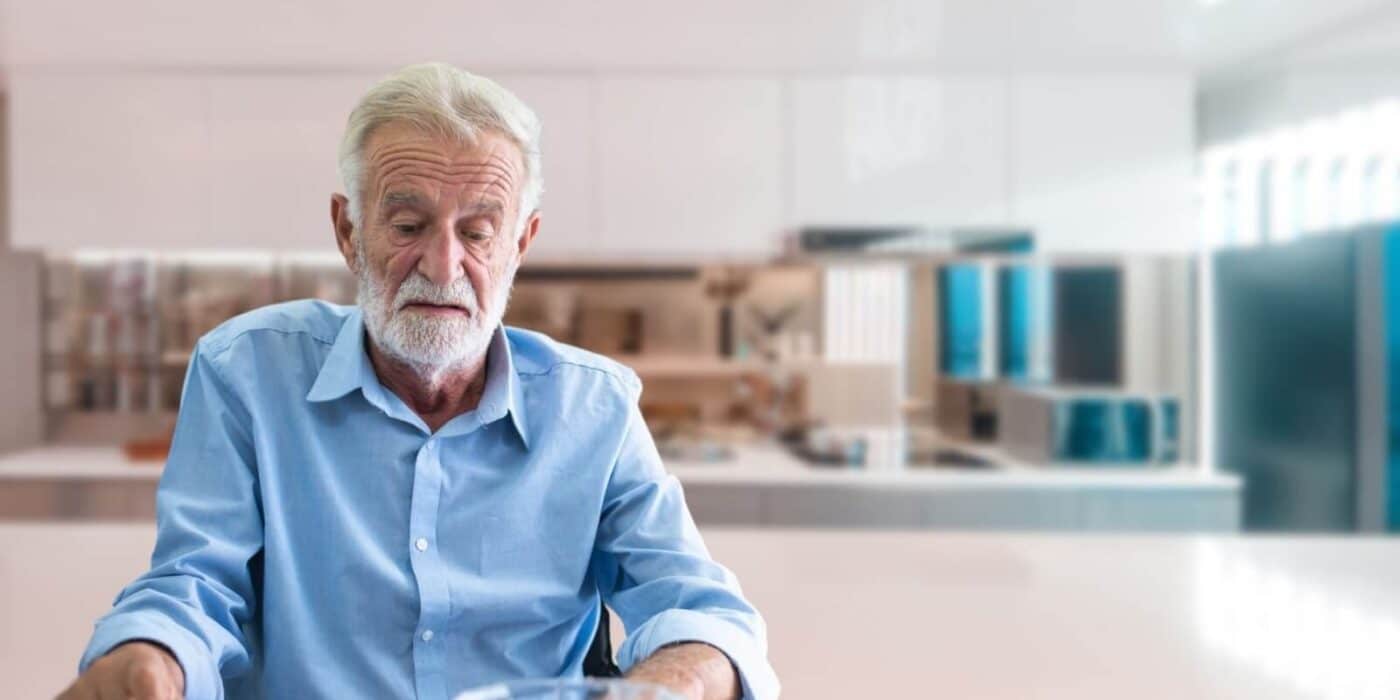 An older adult looks at a bowl of yogurt uninterestedly while sitting alone in their kitchen.