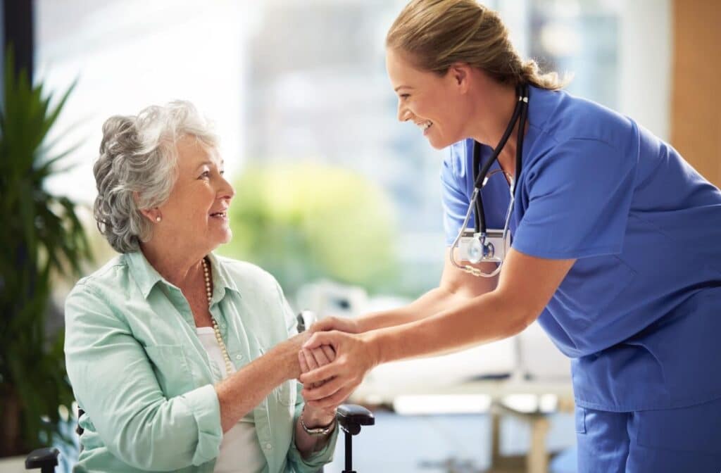 A caregiver smiles while clasping the hands of a seated older adult in front of a sunlit window in a senior living community