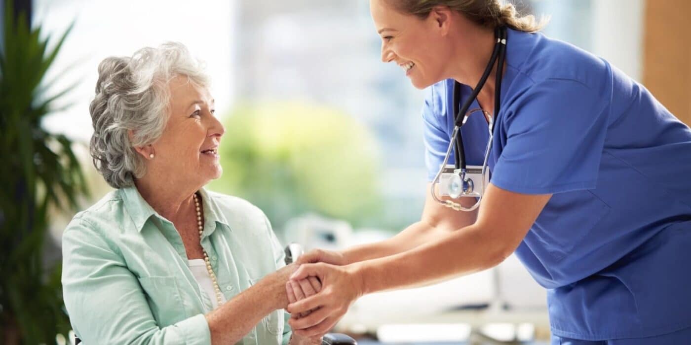 A caregiver smiles while clasping the hands of a seated older adult in front of a sunlit window in a senior living community
