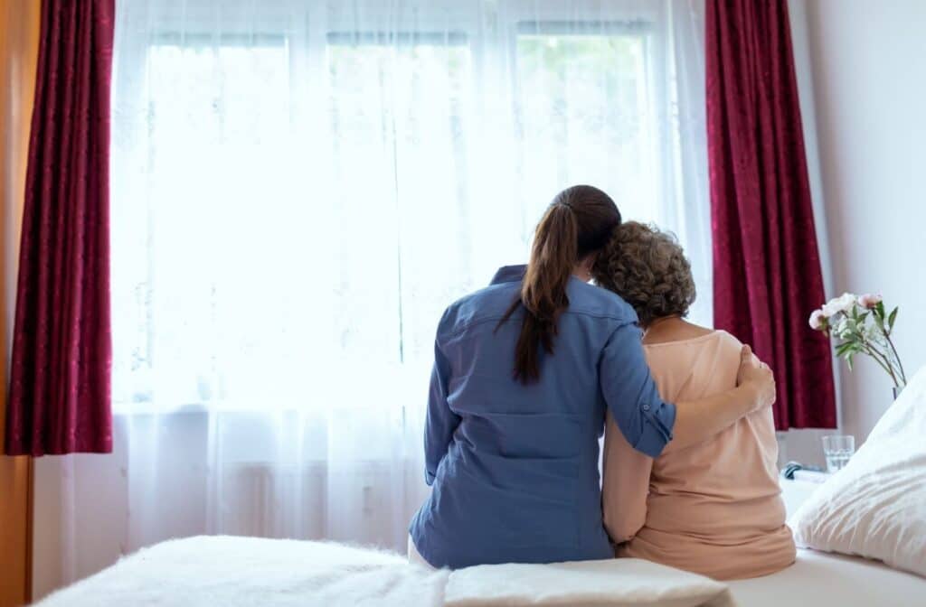 An image from behind of a caregiver hugging an older adult while sitting together on a bed in front of a window in a bedroom.