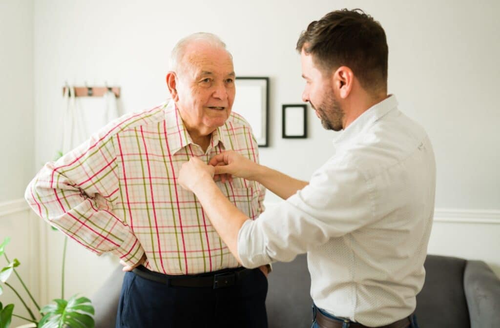 An adult helps their senior parent dress by buttoning a shirt.