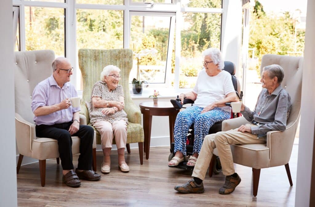A group of seniors sit and talk over cups of coffee in a brightly lit room in their senior living community.