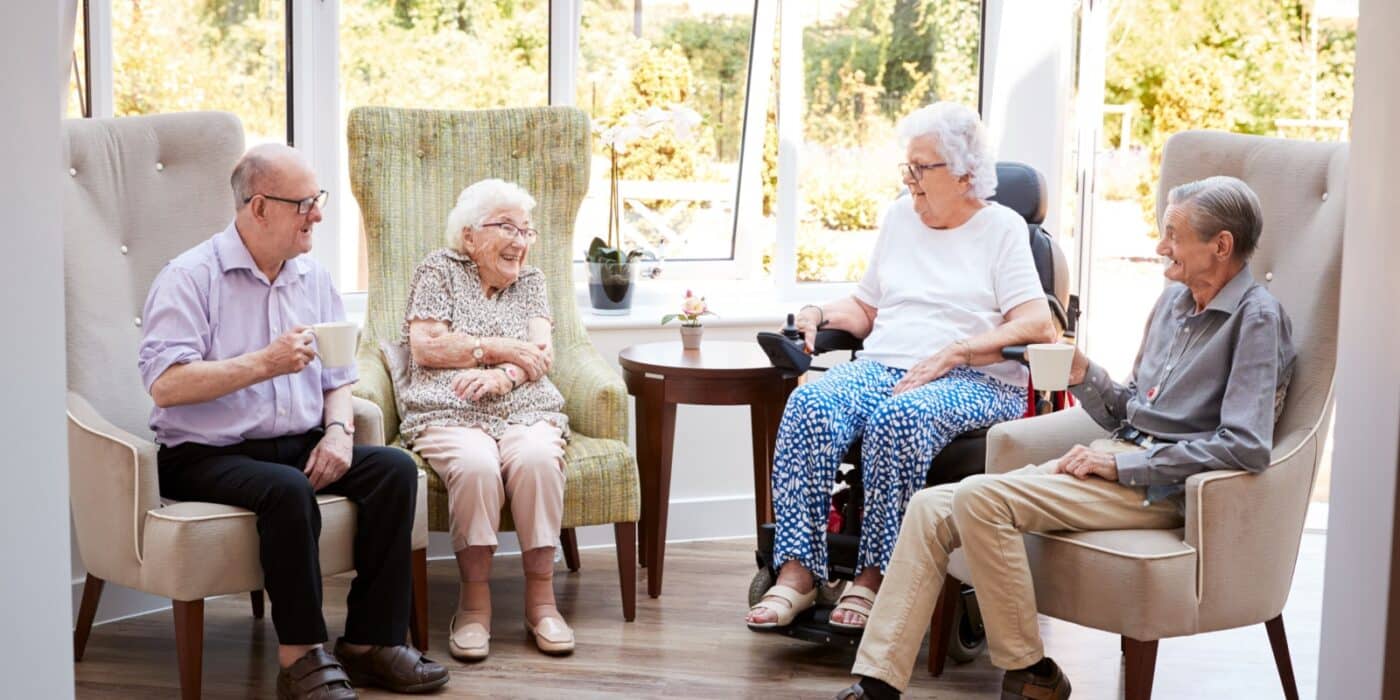 A group of seniors sit and talk over cups of coffee in a brightly lit room in their senior living community.