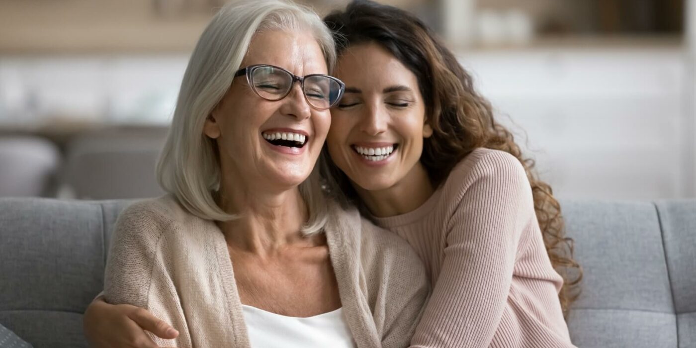 an adult child embraces their senior parent, both smiling warmly in an assisted living apartment.